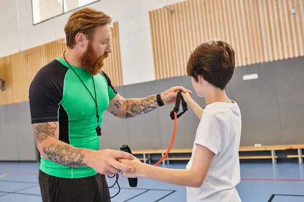 Um homem tatuado ensina um menino a segurar uma corda de salto em uma sala de aula vibrante. — Fotografia de Stock