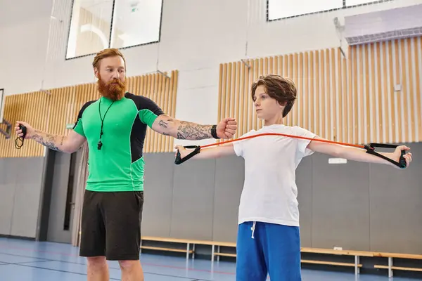 A bearded instructor with tattoos guides a young student through arm stretches in a vibrant school gymnasium. — Stock Photo