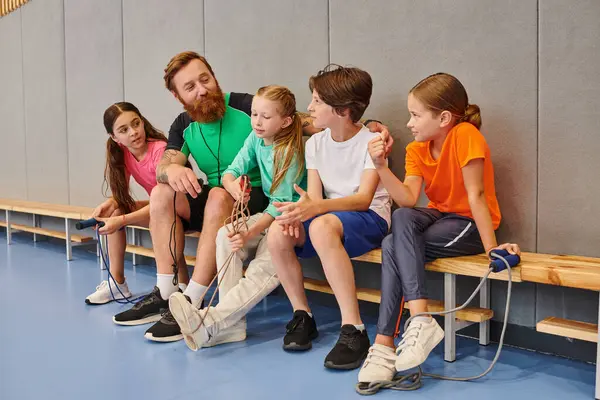 A group of diverse kids sit closely together on a bench, listening attentively to their male teacher in a vibrant gym. — Stock Photo