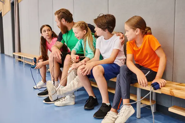 Разнообразная группа маленьких детей, сидящих на скамейке, внимательно слушающих, как их учитель-мужчина передаёт знания в динамичной обстановке класса. — стоковое фото