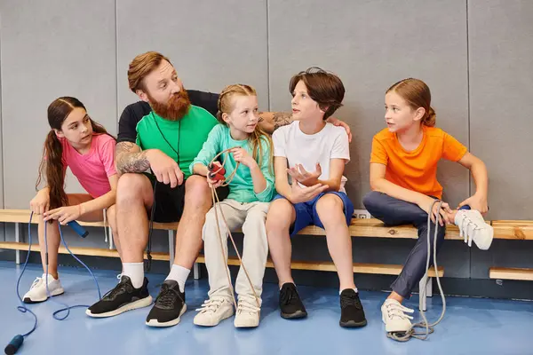 A diverse group of children sitting attentively on a bench, listening to their male teacher in a bright, lively classroom setting. — Stock Photo
