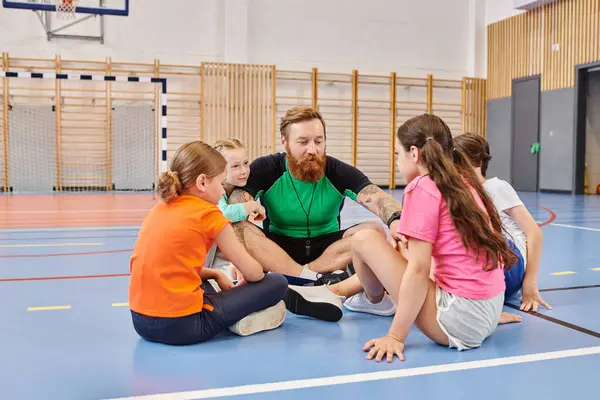 A male teacher sits on the floor surrounded by a diverse group of children in a vibrant school gym, engaged in interactive and educational activities. — Stock Photo