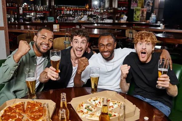 Grupo de amigos interracial emocionados animando con vasos de cerveza en el bar, hombres en despedida de soltero - foto de stock