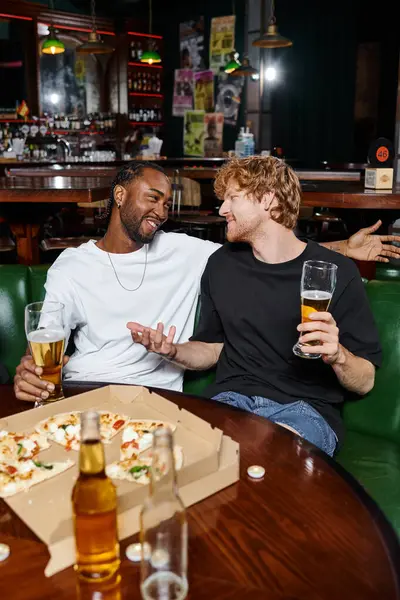 Happy redhead man gesturing while chatting with african american friend over beer and pizza in bar — Stock Photo