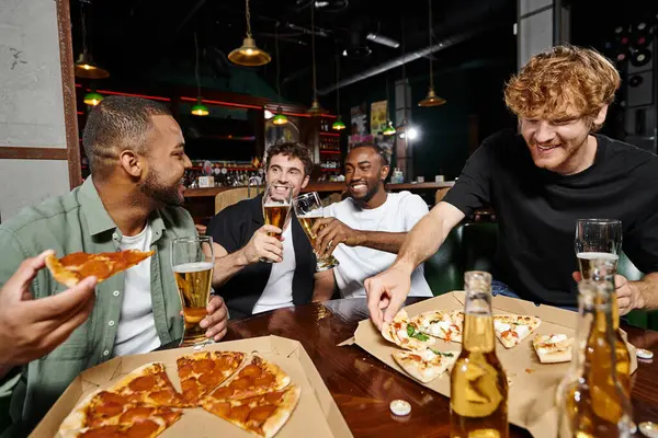 Cuatro alegres amigos multiculturales comiendo pizza y bebiendo cerveza en el bar, hombres en despedida de soltero - foto de stock