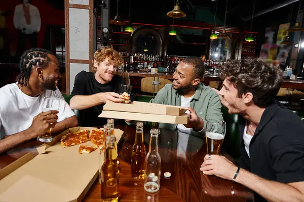 Cuatro amigos multiétnicos felices compartiendo pizza y bebiendo cerveza en el bar, hombres en despedida de soltero - foto de stock