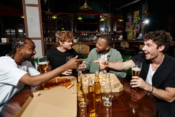 Amigos multiétnicos felizes brincando, gesticulando e conversando sobre pizza com cerveja, homens na despedida de solteiro — Fotografia de Stock