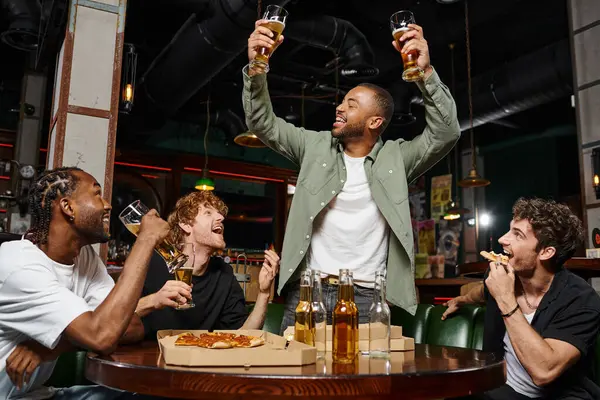Alegre afroamericano hombre levantando dos vasos de cerveza cerca de amigos durante la despedida de soltero en el bar - foto de stock