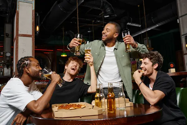 Hombre afroamericano feliz levantando dos vasos de cerveza cerca de amigos durante la despedida de soltero en el bar - foto de stock