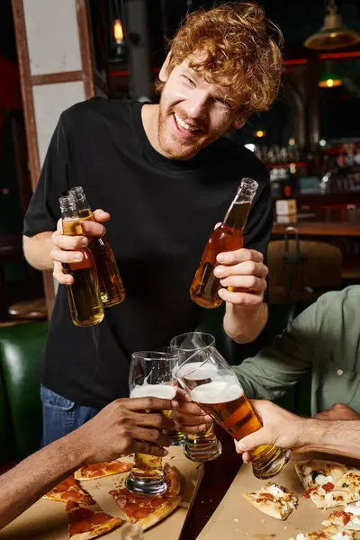 Happy redhead man with curly hair holding bottles with beer near friends toasting glasses in bar — Stock Photo