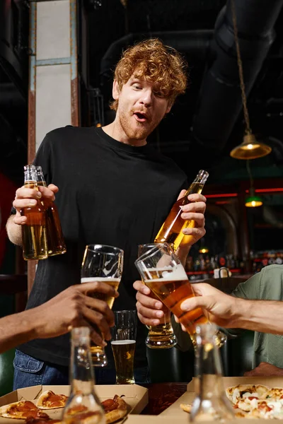 Excited redhead man with curly hair holding bottles with beer near friends toasting glasses in bar — Stock Photo
