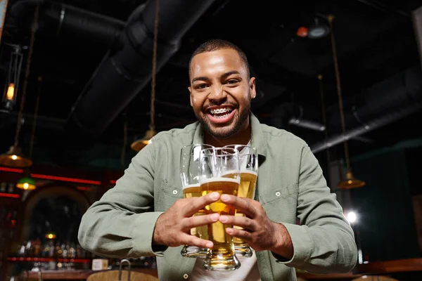 Feliz homem americano africano com aparelho segurando três copos de cerveja no bar, se divertindo nos fins de semana — Fotografia de Stock