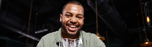 Bandeira de homem americano africano feliz com aparelho segurando três copos no bar, se divertindo — Fotografia de Stock