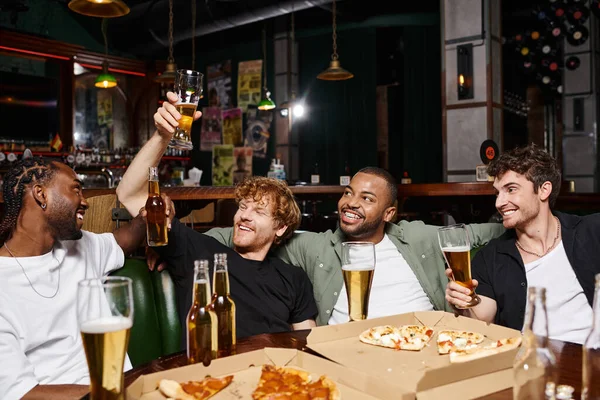 Four happy interracial friends raising bottles and glasses of beer in bar, spending time together — Stock Photo