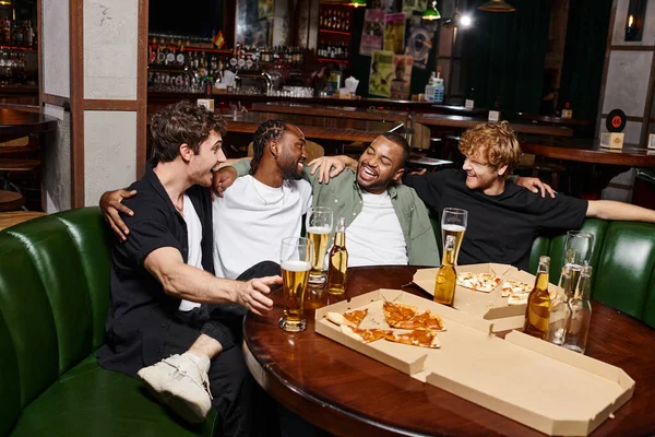 Cuatro felices amigos interracial abrazando y charlando cerca de pizza y cerveza, pasar tiempo en el bar - foto de stock