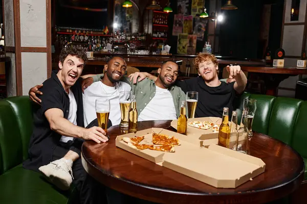 Quattro allegri amici interrazziali che si abbracciano e si siedono vicino a pizza e birra, trascorrendo del tempo al bar — Foto stock
