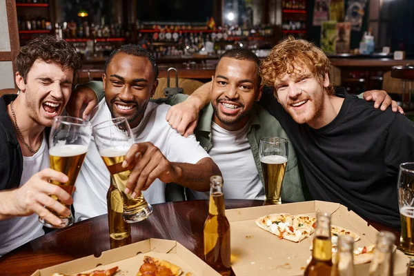 Group of happy multiethnic friends hugging and toasting with glasses of beer, spending time in bar — Stock Photo