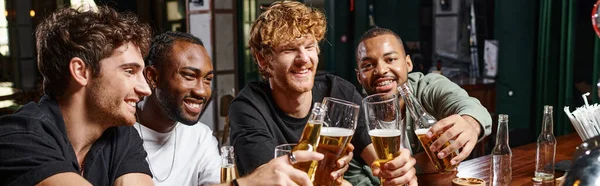 Group of four happy multiethnic male friends toasting with glasses of beer at bar counter, banner — Stock Photo