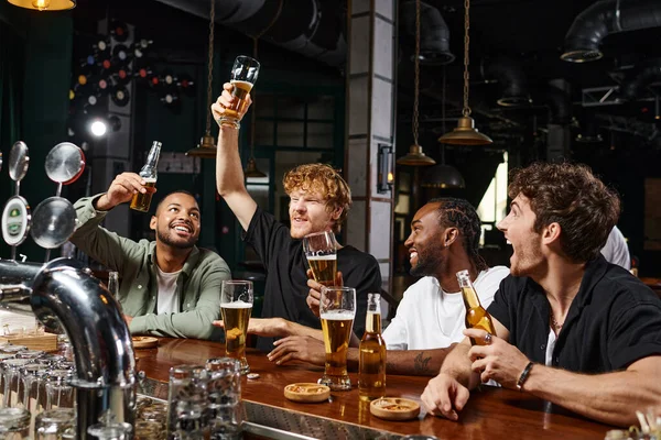Grupo de cuatro amigos masculinos multiétnicos felices levantando vasos de cerveza en el mostrador del bar, despedida de soltero - foto de stock