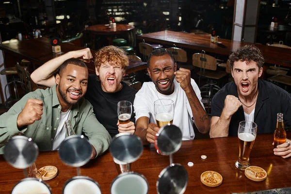 Quatro excitados fãs de futebol multiétnico segurando copos de cerveja e torcida, amigos do sexo masculino no bar — Fotografia de Stock