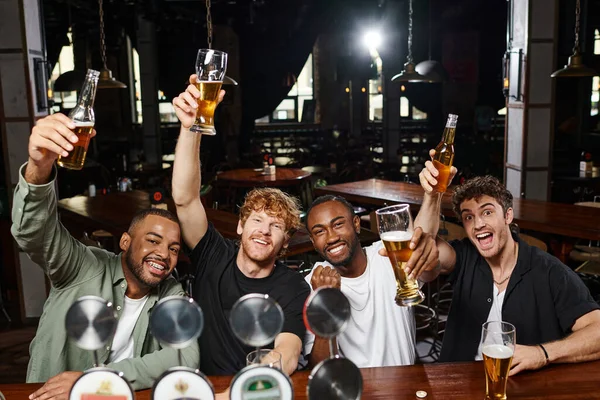 Quatre hommes multiethniques excités lever des verres de bière pendant la fête de célibataire, amis masculins dans le bar — Photo de stock