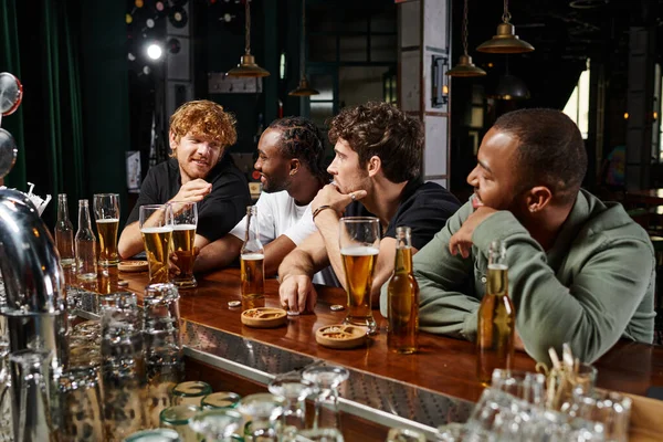 Grupo de hombres multiculturales pasar tiempo juntos, charlando y bebiendo cerveza, amigos varones en el bar - foto de stock