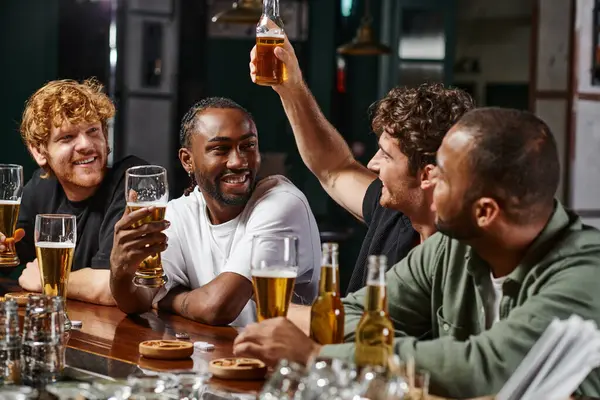 Fröhliche multikulturelle Männer, die ihren Freund dabei beobachten, wie er ein Glas Bier anhebt, während er Zeit miteinander in der Bar verbringt — Stockfoto