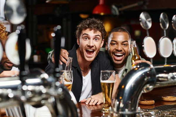 Animado interracial amigos abraçando e segurando cerveja, passar tempo no bar, amizade masculina — Fotografia de Stock