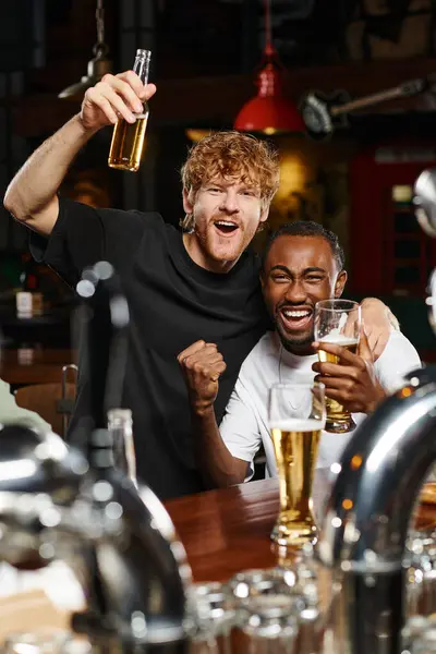 Возбужденные межрасовые друзья обнимаются и поднимают бокалы пива, проводя время в баре — стоковое фото