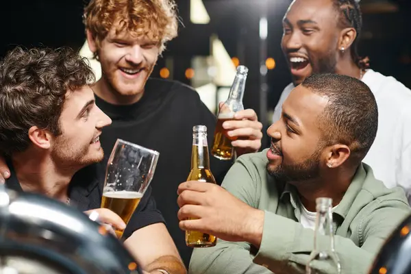 Vier fröhliche multiethnische Freunde lächeln und prosten Biergläser zu, während sie Zeit in der Bar verbringen — Stockfoto