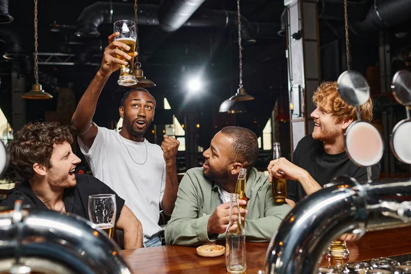 Hombre afroamericano asombrado levantando un vaso de cerveza cerca de amigos alegres en el mostrador del bar, vida nocturna - foto de stock