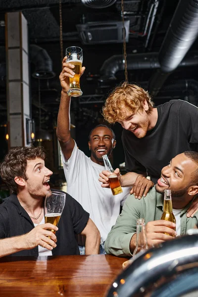 Hombre afroamericano excitado levantando un vaso de cerveza cerca de amigos alegres en el mostrador del bar, la vida nocturna - foto de stock