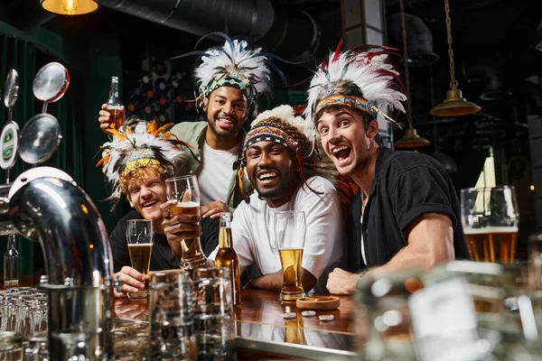Amigos masculinos multiétnicos emocionados en ropa de cabeza con plumas tostando cerveza y pasar tiempo en el bar - foto de stock