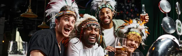 Pancarta, amigos masculinos multiétnicos en ropa de cabeza con plumas tostadas cerveza y pasar tiempo en el bar - foto de stock
