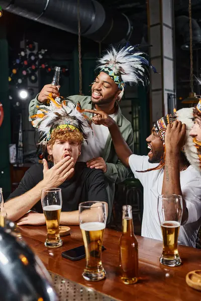 Hombres multiculturales felices en la cabeza con plumas sosteniendo vasos de cerveza y divirtiéndose en el bar - foto de stock