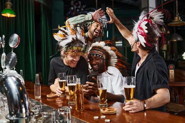 Щасливі мультикультурні чоловіки в головній убір з пір'ям, які спостерігають за футбольним матчем на смартфоні в барі — стокове фото