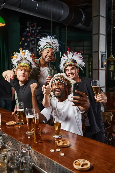 Homens multiétnicos alegres em chapéus com penas tirando selfie no smartphone durante a despedida de solteiro — Fotografia de Stock