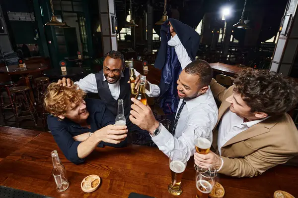 Grupo de cuatro colegas felices en ropa formal bromeando y bebiendo cerveza en el bar, divirtiéndose después del trabajo - foto de stock