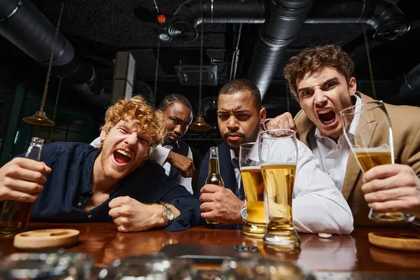 Colegas multiculturales emocionales en ropa formal sosteniendo cerveza y gritando en el bar, los hombres después del trabajo - foto de stock