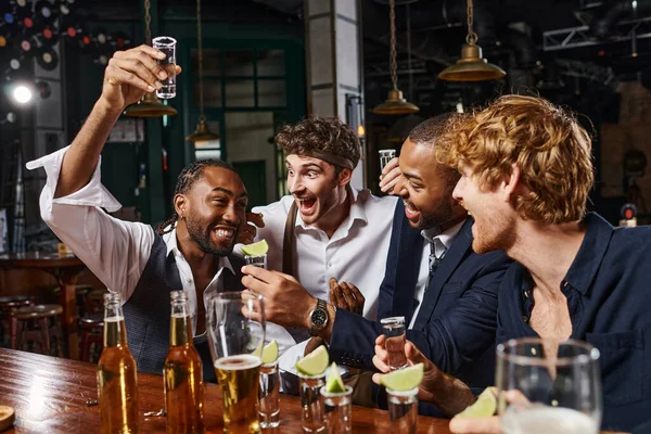 Feliz africano americano hombre levantando disparo de tequila cerca emocionado amigos durante despedida de soltero en bar - foto de stock
