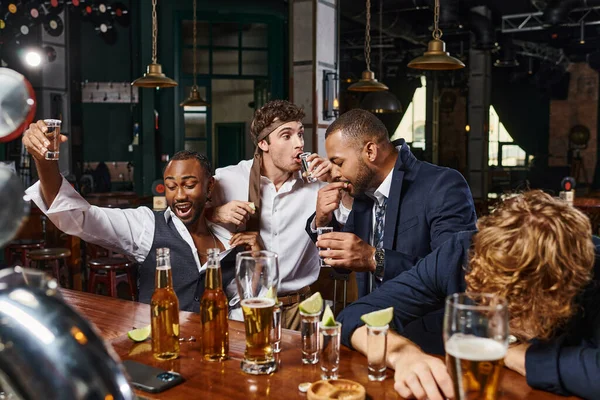 Foto espontânea de homens multiculturais engraçados e bêbados em uso formal bebendo tequila no bar após o trabalho — Fotografia de Stock