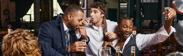 Pancarta de hombres divertidos y borrachos multiculturales en ropa formal beber tequila en el bar después del trabajo - foto de stock