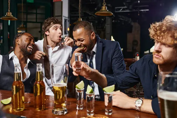 Candid photo of funny and drunk interracial men in formal wear drinking tequila in bar after work — Stock Photo