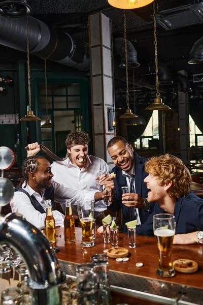 Candid photo of happy and drunk interracial men in formal wear drinking tequila in bar after work — Stock Photo