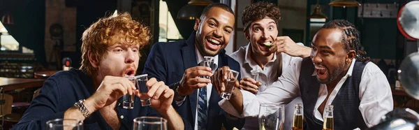 Pancarta de cuatro hombres interracial felices y borrachos en ropa formal bebiendo tequila en el bar después del trabajo - foto de stock