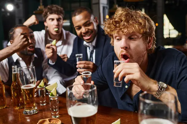 Enfoque en el hombre pelirrojo beber tequila tiro cerca interracial borracho amigos en el bar después del trabajo - foto de stock