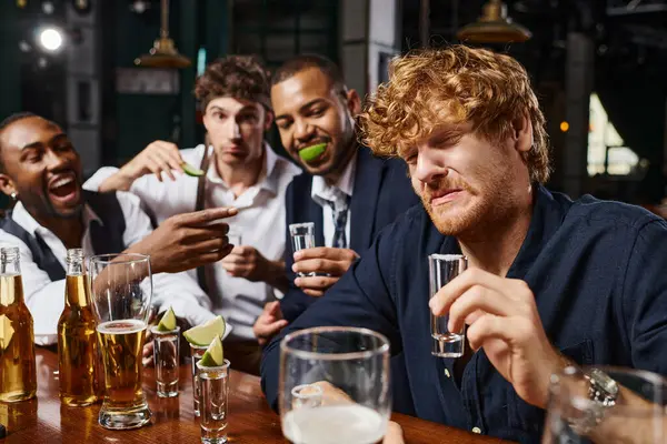 Centrarse en el hombre pelirrojo mueca mientras bebe tequila tiro cerca de amigos interracial después del trabajo - foto de stock