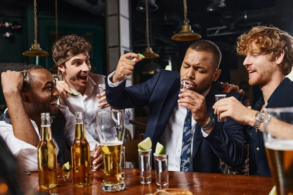 Happy interracial men looking at african american friend drinking tequila shot and holding lime — Stock Photo