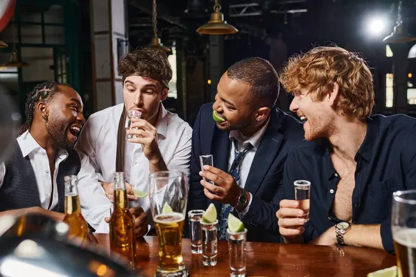 Happy interracial men looking at drunk friend with tie on head drinking tequila shot in bar — Stock Photo