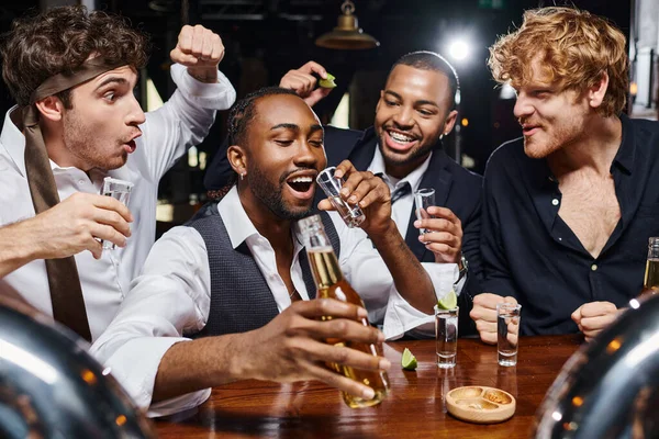 Feliz interracial homens torcendo perto de afro americano amigo beber tequila tiro e segurando cerveja — Fotografia de Stock